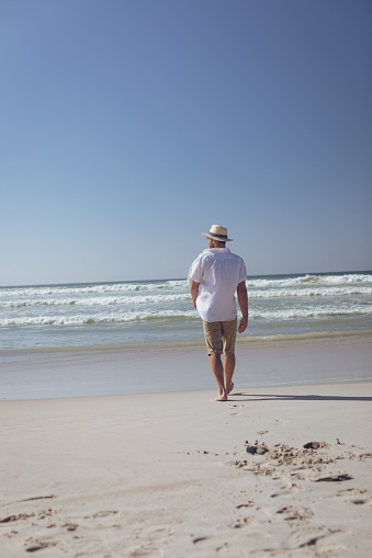 Rear view of young Caucasian man walking at beach on a sunny day. He wears a hat