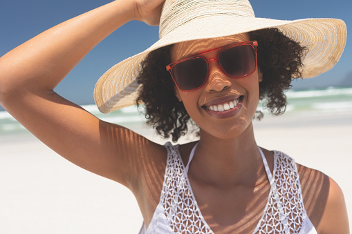 Portrait of beautiful young mixed-race woman smiling and looking at camera standing at beach on a sunny day. She wears hat and red sunglasses