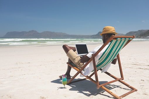 Side view of African-American man using laptop while sitting at sun lounger at beach on a sunny day. He wears hat