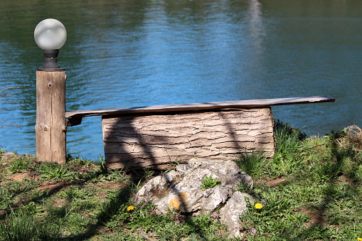 Wooden log used as improvised bench in shade of large tree next to round white backyard outdoor light mounted on tree stump and large rock on grass covered river bank overlooking clear water on warm sunny spring day