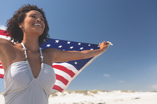 Portrait of happy Mixed-race woman holding american flag at beach on a sunny day. Woman is wearing swimsuit
