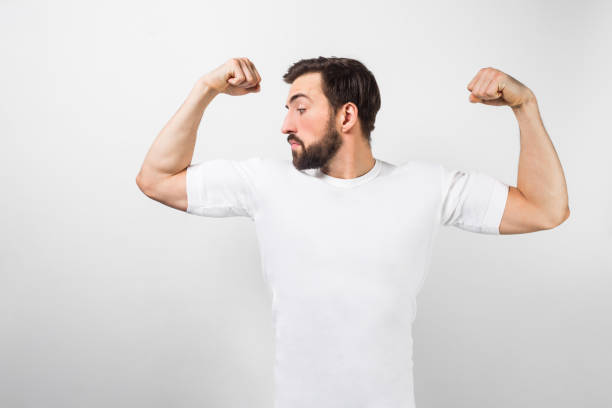 istock A handsome confident young man standing and showing big muscles on his hands. He is looking at one of them and very proud of them. He is wearing a white t-shirt, isolated on white background. 1140112346