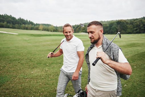 Two stylish men holding bags with clubs and walking on golf course