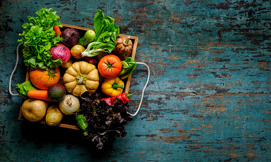 Many colorful contrast color salad vegetables, shot from above, sitting in an old wooden vegetable tray on an abstract blue/turquoise wood table background with atmospheric lighting, with atmospheric lighting, good copy space to the right of the image.