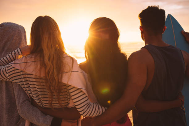Rear view of a group of friends standing together on beach holding each other. Man standing with his women friends at the beach looking at the sunset holding a surfboard.