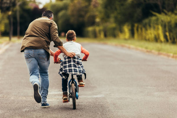 Man helping his kid in learning to ride a bicycle Rear view of a boy riding a bicycle while his father runs along holding the kid. Father teaching his son to ride a bicycle. riding stock pictures, royalty-free photos & images