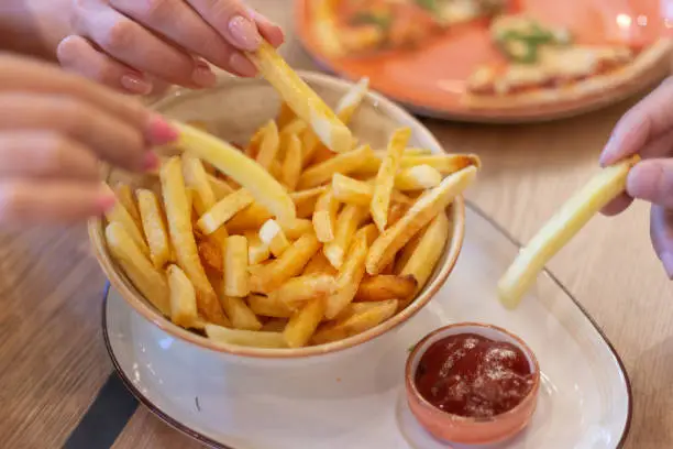 Hands of three young people sharing bowl of french fries and dipping them into ketсhup before eating in restaurant