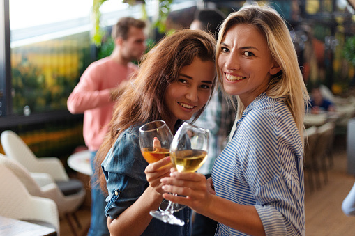 Two beautiful women posing with glasses of wine and smiling at camera at party in bar