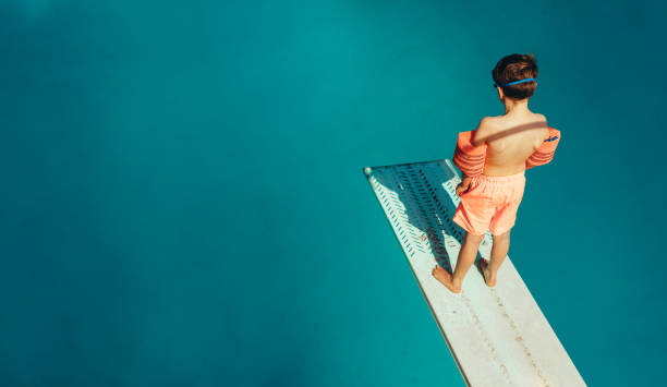 Boy standing on a diving board Top view of boy standing on spring board learning to dive during swimming class on a summer day. Boy learning swimming at outdoor pool. diving board stock pictures, royalty-free photos & images