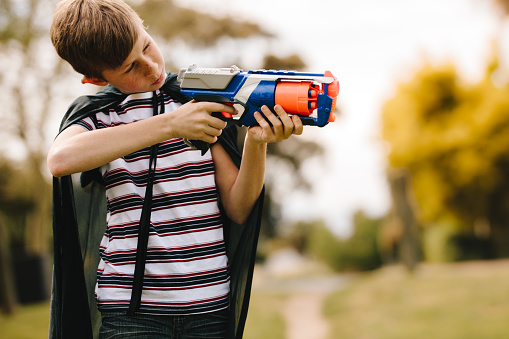 Young boys wearing a cape playing with a toy gun outdoors. Boy with cape pretending to a superhero.