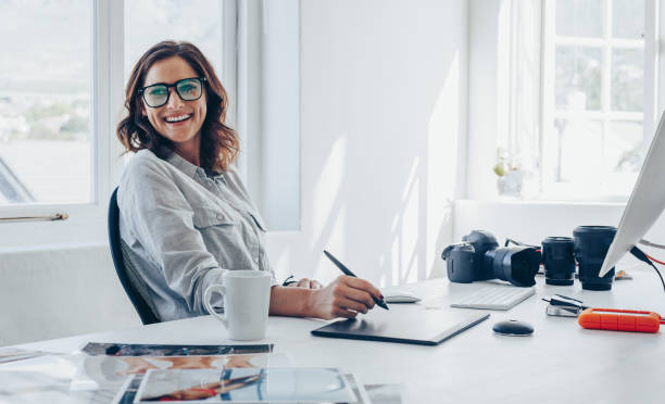 Professional photographer at her office desk Professional photographer sitting at her office desk looking away and smiling. Woman in office with digital graphic tablet and drawing pen. graphic designer photos stock pictures, royalty-free photos & images