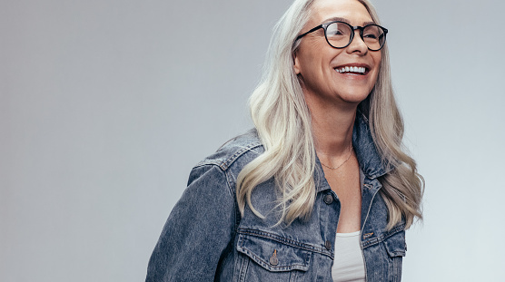Cheerful senior woman in casuals looking away and smiling over grey background. Happy mature woman in denim shirt and eyeglasses.