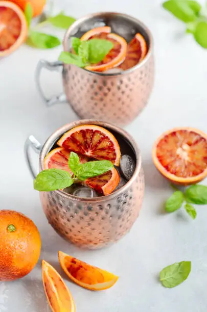 Blood orange Moscow mule alcohol cocktail with fresh mint leaves and ice in copper mugs on a gray concrete background, selective focus.