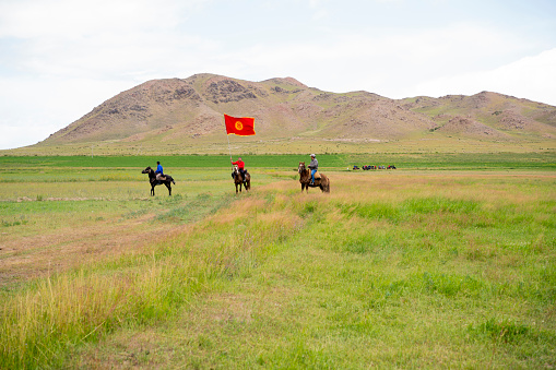 Bokonbayevo, Kyrgyzstan - Avgust 12, 2017: Three Kyrgyz horsemen with flag in middle of them, riding horses in steppe during nomad traditional festival - Kyzil Oi (Birds or Prey Festival) not far from small town Bokonbayevo in the Southern shore of Issyk-Kul lake.