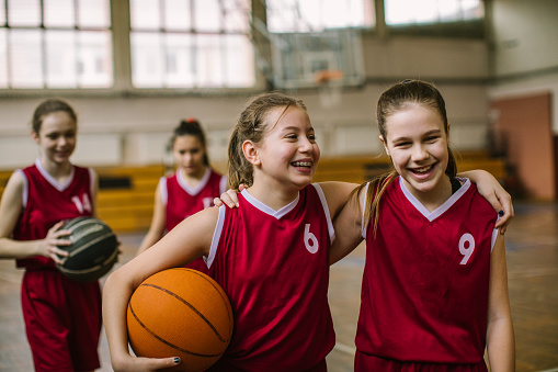 A shot of a female teenage athlete sitting on the floor during half time of a basketball game. She is smiling and looking into the camera. She is socialising with her team mates with a basketball by her side.