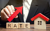 A man holds a red arrow up above the word Rate and a wooden house. The concept of raising interest rates on mortgages. The increase in property tax rates. Real estate capitalization. Insurance.