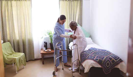 Front view of mixed race female nurse helping senior mixed race female patient to stand with walker at nursing home