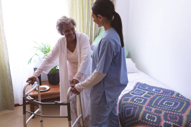 Female nurse helping senior female patient to stand with walker Front view of mixed race female nurse helping senior African American female patient to stand with walker at nursing home sheltering photos stock pictures, royalty-free photos & images