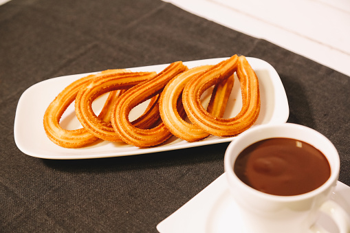 spanish fritters served on a wooden table with a gray tablecloth and a cup of hot chocolate