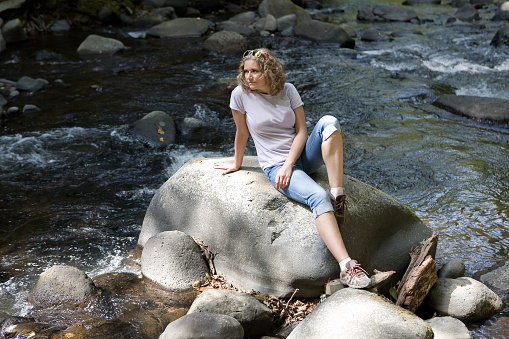 A beautiful young woman is sitting on a stone next to a mountain stream. Human and nature.