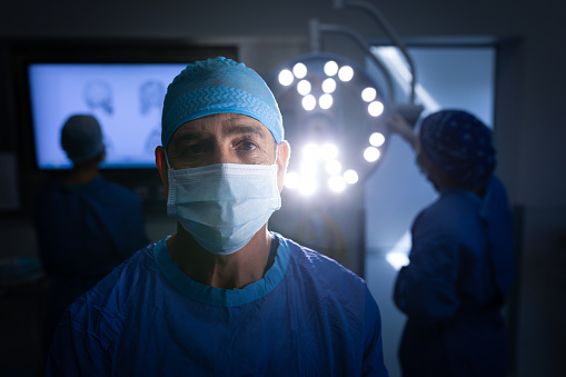 Portrait of a Caucasian male surgeon wearing an operation mask and looking at the camera in the operation theater with coworkers working behind