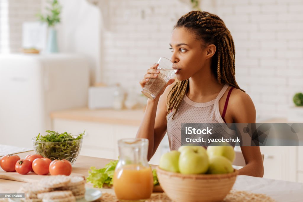Slim and fit woman drinking water before having breakfast Water before breakfast. Slim and fit woman with many little braids drinking water before having breakfast Water Stock Photo