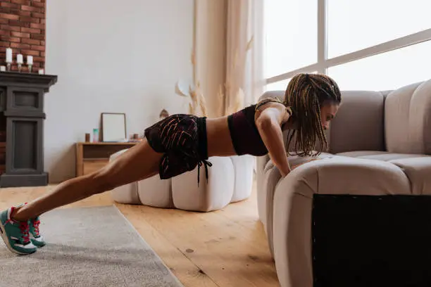 Doing pushups. Active fit woman wearing shorts and top doing pushups near sofa at home