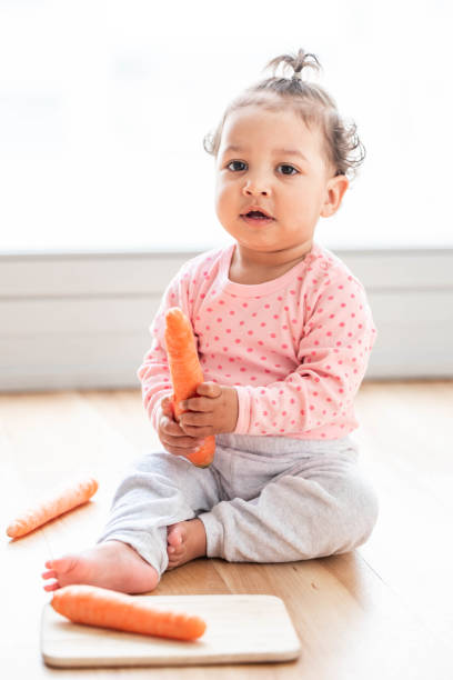 niedliches mädchen auf dem boden sitzt und mit einer karotte spielt - baby carrot stock-fotos und bilder