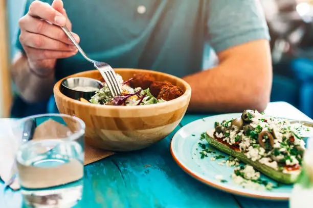 Shot of a young man eating fresh vegan salad with chickpeas balls and sesame sauce on rustic wooden table