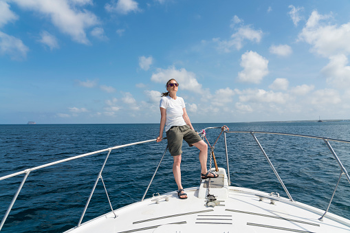 Mature woman standing on the edge of a boat in the middle of blue sea. Blue sky in background on sunny day. She wears t-shirts and shorts, front view.   Galapagos
