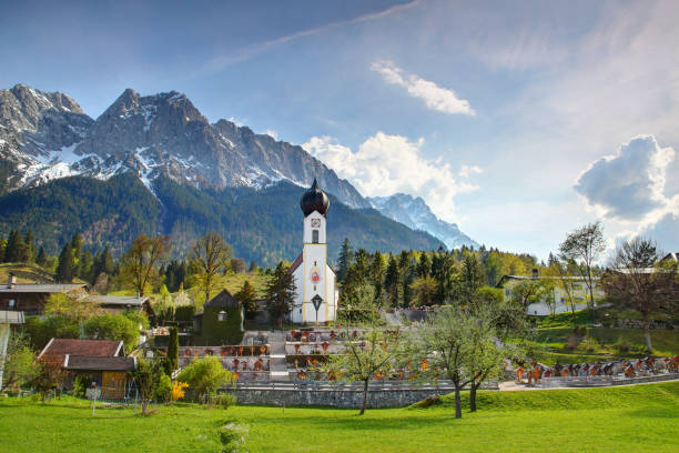 piccola chiesa bianca st johannes, cimitero e case in primavera sotto il cielo azzurro con soleggiate cime di waxenstein e zugspitze sullo sfondo, wetterstein northern limestone alps grainau garmisch-partenkirchen bayern germania europa - grainau foto e immagini stock