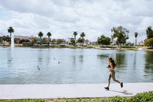 A Latina woman runs and performs bodyweight exercises at MacArthur Park in Los Angeles, California on a bright sunny day.  She jogs along the edge of MacArthur Lake.