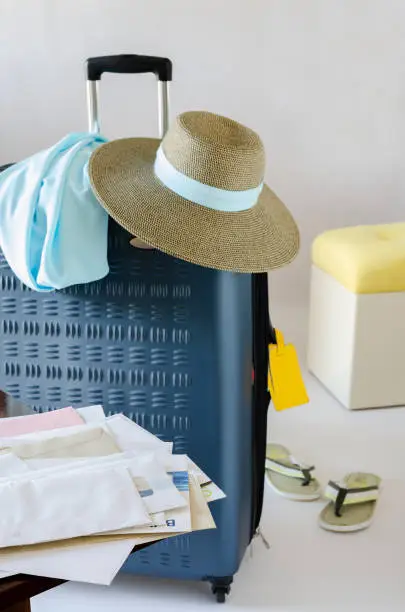 Big blue rectangular suitcase for vacation travel with brown sunhat and blue neckpiece on it; a pair of grey beach flip-flops; yellow pouf; heap of mail; gray background