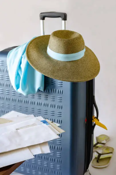 Big blue rectangular suitcase for vacation travel with brown sunhat and blue neckpiece on it; a pair of grey beach flip-flops; heap of mail; gray background