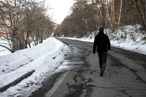 A man walking down an empty Winter road, lined by snow.