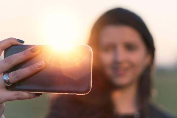 pretty young girl smiling and taking selfies, solo traveler, taking pictures, one young woman tourist using smartphone, enjoying the sunset light, portrait, back lit, travel, exploration, tourism, outdoors, generation z, springtime - back lit women one person spring imagens e fotografias de stock