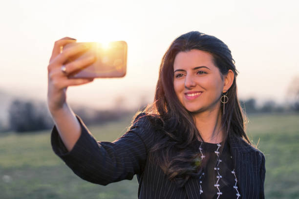 pretty young girl smiling and taking selfies, solo traveler, taking pictures, one young woman tourist using smartphone, enjoying the sunset light, portrait, back lit, travel, exploration, tourism, outdoors, generation z, springtime - back lit women one person spring imagens e fotografias de stock