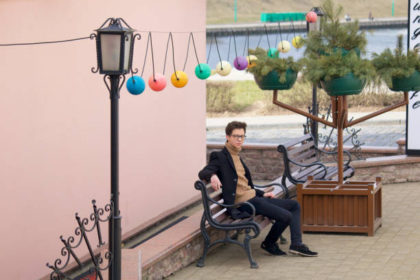 istock A young man with a pensive face is sitting on a bench. Dressed in a black light overcoat, black-rimmed glasses. 1140312067