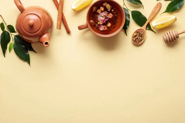 Traditional tea ceremony accessories, teapot and teacup with cinnamon sticks, spoon of dried tea, green leaves, honey dipper on yellow background, top border, flat lay composition, tea time concept