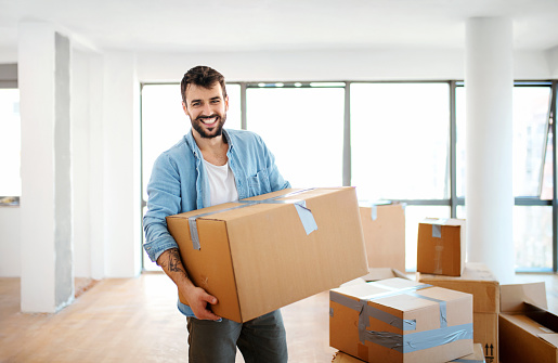 Closeup front view of a smiling young man carrying boxes as he moves into his new apartment.