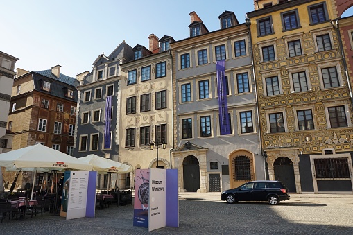 Warsaw, Poland - 22 March 2019: View of a street cafe and the Museum of Warsaw in a corner of Warsaw's Old Town Market Square in afternoon sunshine. The sun is just beginning to cast shadows across the frontage. A couple of advertising boards add colour.