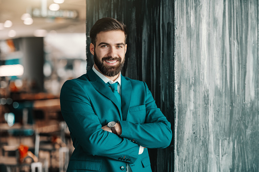 Smiling bearded ambitious Caucasian businessman in suit standing with arms crossed and leaning on pillar in cafe. Mistakes are proof that you are trying.