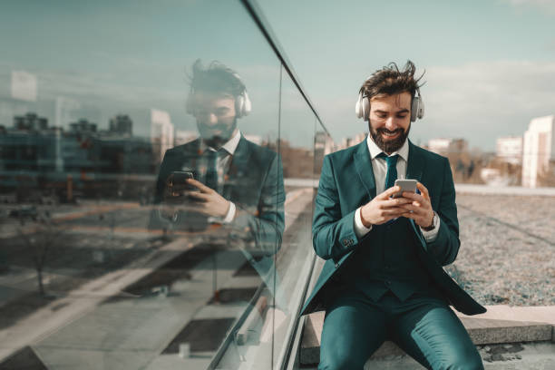 sorridente uomo d'affari caucasico barbuto di successo in abbigliamento formale ascoltando musica e usando lo smartphone mentre è seduto sul tetto. - roof men business city foto e immagini stock