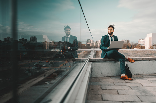 Young smiling cheerful Caucasian bearded businessman in formal wear using laptop while sitting on rooftop. At first they will ask you why you are doing it, later they will ask you how you did it.