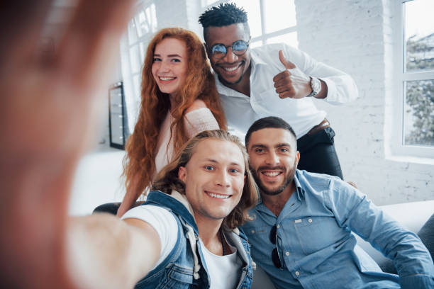 istock Guy shows thumb up. Cheerful young friends taking selfies on sofa and white interior 1140328648