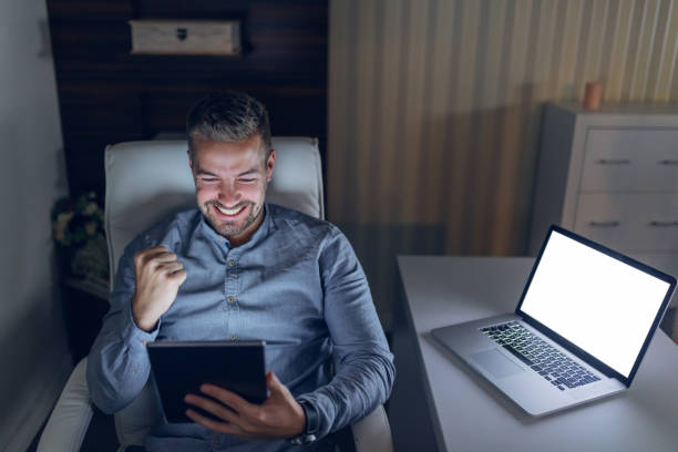 istock Happy young handsome freelancer holding tablet and cheering for nicely done job. On desk laptop. Late night work concept. 1140333235