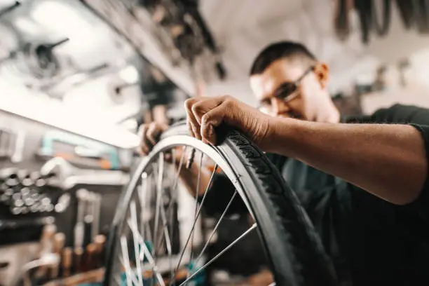 Photo of Caucasian dedicated man putting tyre on bicycle wheel while standing in workshop.