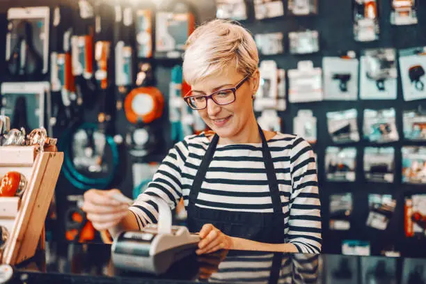 Photo of Smiling Caucasian female worker with short blonde hair and eyeglasses using cash register while standing in bicycle store.