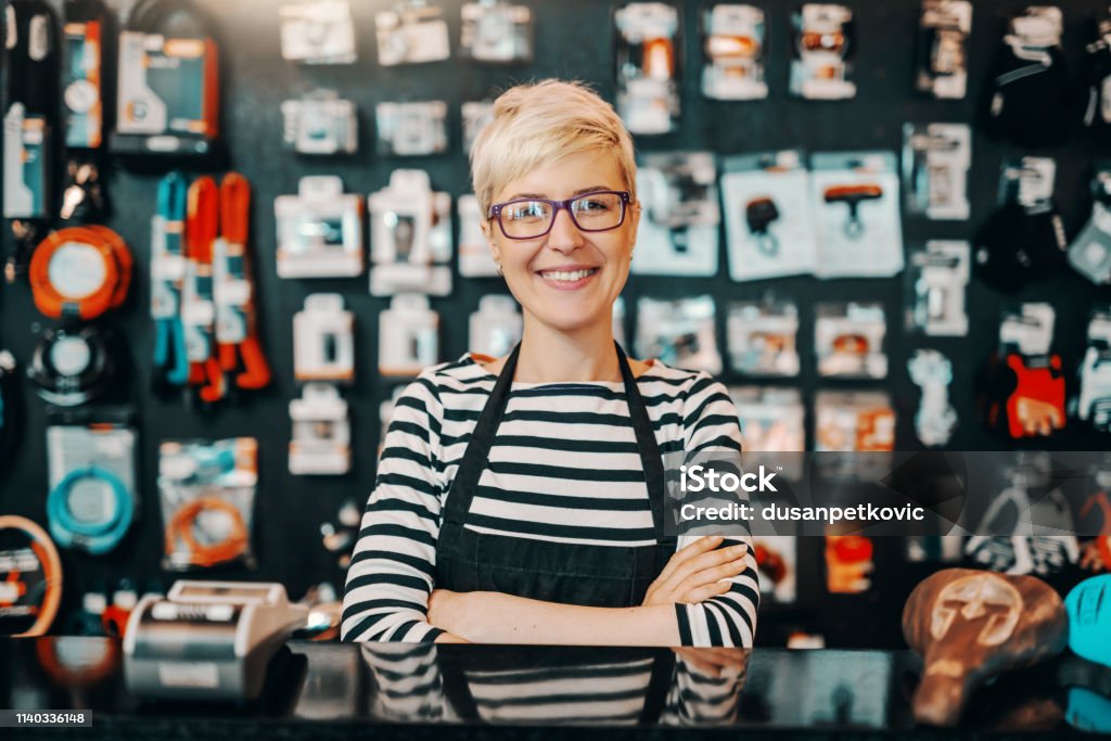 Portrait of beautiful smiling Caucasian female worker with short blonde hair standing in bicycle shop with arms crossed. Store Stock Photo