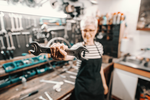 Portrait of beautiful Caucasian female worker holding wrench while standing in bicycle workshop. Selective focus on wrench.